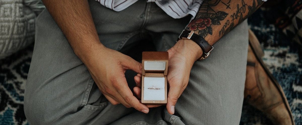 sediaci muž s tetovaním na predlaktí držiaci v rukách krabičku so zásnubným prsteňom